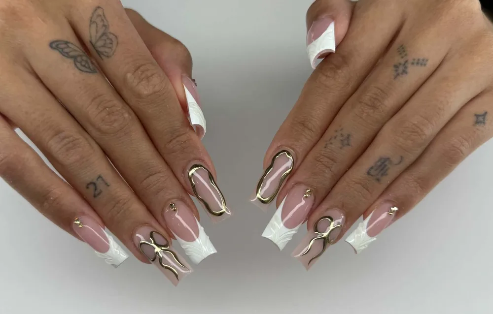 Long square nails with textured white French tips and raised gold metallic swirls over a nude base