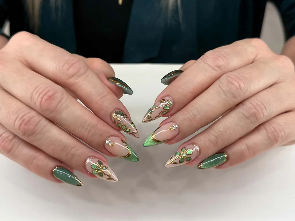 Stiletto nails in nude with emerald cat-eye tips, hand-painted green and gold foliage, and small crystals